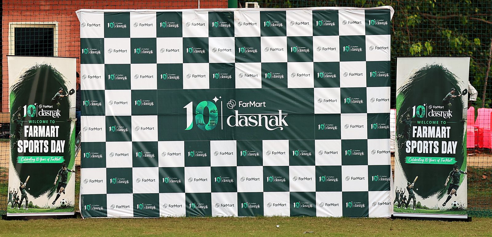 A green and white checkered step-and-repeat backdrop alternating the FarMart and dashak logos, with the large lockup in the centre, flanked by two Sports Day roll-ups on a cricket ground.