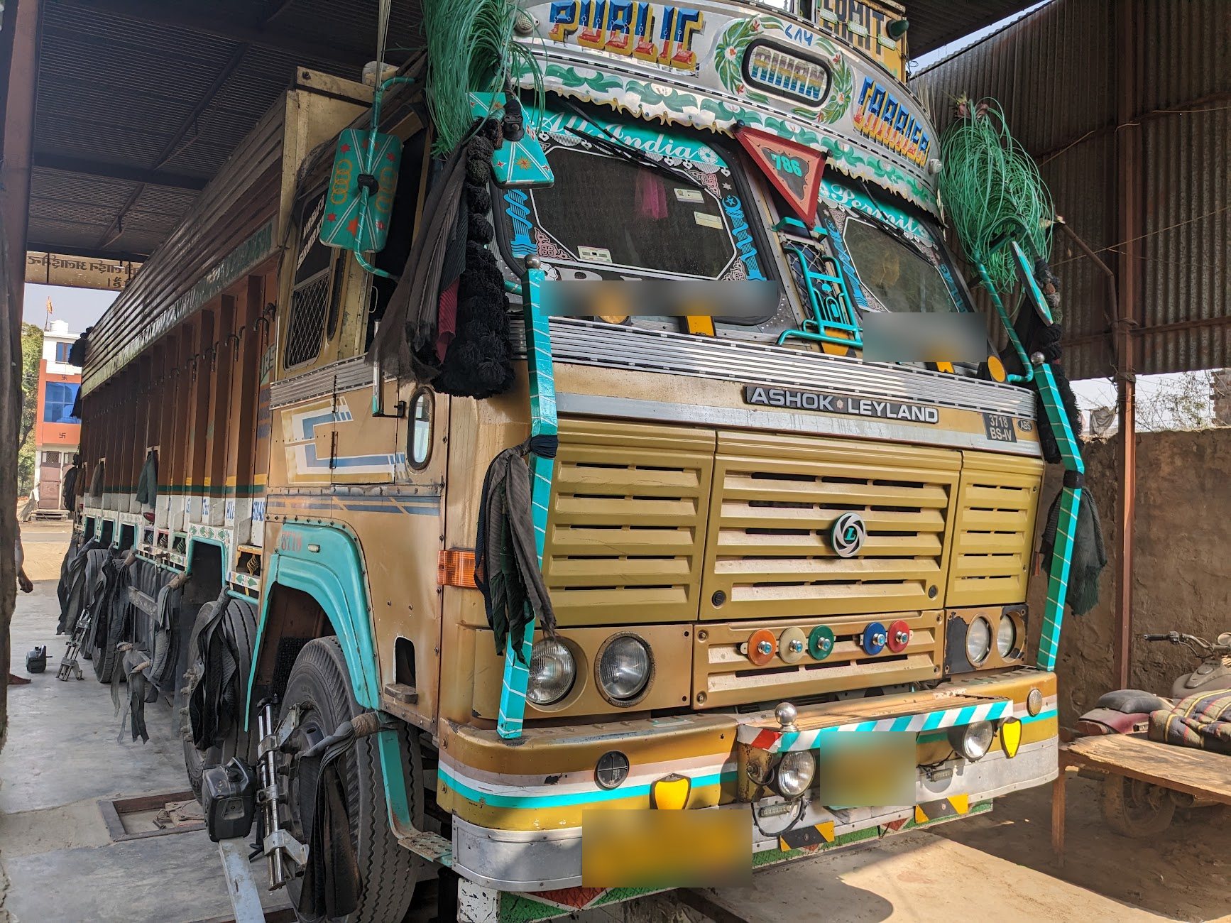 A decorated Ashok Leyland goods carrier truck parked under a shed. Its registration numbers are blurred.